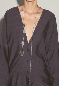 Lange silberne Halskette mit flachen, ovalen Anhängern in Grautönen und Schwarz, getragen über einem dunklen, strukturierten V-Ausschnitt-Shirt.