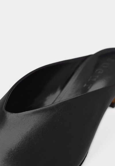 Κοντινή λήψη μιας μαύρης δερμάτινης μύτης mule παπουτσιού με χαμηλό τακούνι και λεία υφή σε λευκό φόντο.