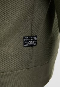 Hanorac verde măsliniu cu un model texturat în zigzag și o etichetă neagră cu text alb în partea inferioară stângă.