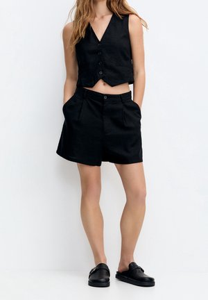 Femme portant un gilet noir sans manches boutonné, un short noir taille haute avec poches, et des mocassins noirs sans lacets, debout devant un fond blanc.