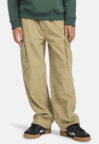 Beige Cargo-Hosen mit entspannter Passform, seitlichen Taschen und einer weichen Baumwollmischung. Kombiniert mit schwarzen Sneakern, die eine strukturierte Oberfläche zeigen.