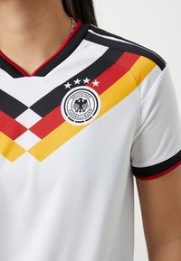 Maillot de football blanc avec des rayures diagonales noires, rouges et jaunes et l'emblème du Deutscher Fussball-Bund avec quatre étoiles sur la poitrine.