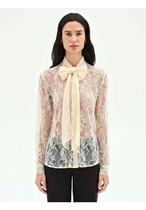 Donna che indossa una camicetta in pizzo beige trasparente con maniche lunghe e un grande fiocco al collo, abbinata a pantaloni neri, in piedi contro uno sfondo bianco.