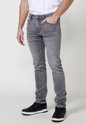 Jeans de mezclilla gris ajustados con un acabado desgastado, diseño de cinco bolsillos y detalles de costura, combinados con zapatillas negras.
