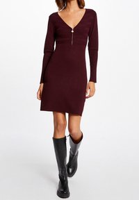 Burgundské pletené šaty s dlouhými rukávy, V-výstřihem a detailním předním zipem. Vyrobené z elastického materiálu, hladká textura, délka nad kolena.