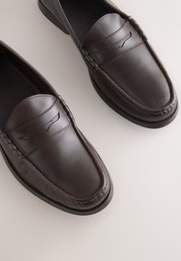 Paire de mocassins en cuir brun foncé avec des détails cousus et une fente pour pièce sur un fond clair neutre.