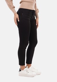Donna che indossa jeans skinny neri, un maglione beige e sneakers bianche con dettagli neri sul tacco, in piedi con le mani appoggiate sulle cosce.