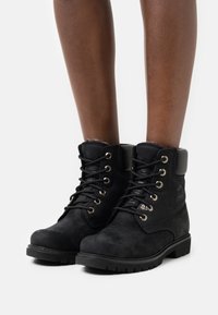 Botines negros de material parecido a ante, con un tacón texturizado, ojales de metal y un cuello acolchado. Suela gruesa con ranuras.