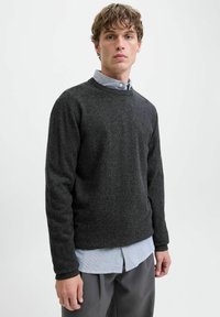 Jeune homme aux cheveux bouclés portant un sweat en col rond gris foncé par-dessus une chemise rayée bleu clair et un pantalon gris, sur un fond uni.
