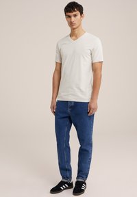 Gai pelēks V-veida kakla T-krekls ar īsām piedurknēm, apvienots ar ziliem taisnajiem džinsiem un melniem sporta apaviem ar baltām svītrām.