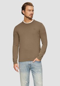 Rjav pleteni pulover s okroglim izrezom in rebrastimi manšetami, oblečen preko bele srajce, kombiniran z svetloblue raztrganimi kavbojkami.