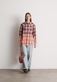 Camisa de botão em padrão xadrez rosa e roxo com dois bolsos no peito; combinada com jeans azul claro e uma bolsa rosa. Sapatos estampados marrons.