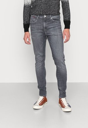 Mężczyzna w ciemnoszarych dopasowanych jeansach, wysokich trampkach z brązowymi i białymi elementami oraz czarno-białym swetrze w cętki.