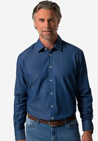 Camicia da uomo a maniche lunghe in tessuto denim blu scuro con colletto, chiusura a bottoni e dettagli in bottoni argentati. Abbinata a jeans chiari.