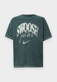 T-shirt oversize di un verde scuro, realizzato in morbido cotone. Presenta una grande stampa grafica con "SWOOSH" e "just do it" in bianco. Design classico con colletto a girocollo.