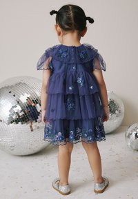 Vestido en azul marino con volantes, mangas abullonadas transparentes, detalles florales bordados y lentejuelas. Combinado con zapatos plateados con brillo.