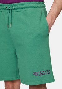 Groene sweatshorts met een hand in de rechterzak en paarse geborduurde "R.S.V.P." tekst op het linkerbeen.
