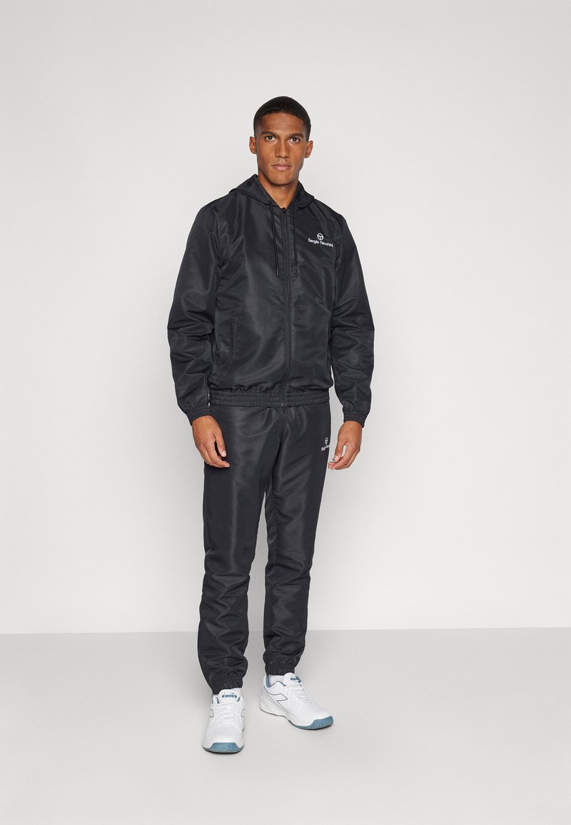 Black nylon track jacket and pants with elastic cuffs, featuring a small white logo on the chest and pants. Model wearing white sneakers.