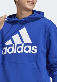 Hoodie in cotone blu con un grande logo bianco "adidas" sul fronte, dotato di cappuccio con coulisse e tasca frontale. Texture liscia.