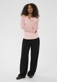 Roze top met lange mouwen en gestructureerde mouwen, gecombineerd met losse zwarte broek. Model staat tegen een neutrale achtergrond.