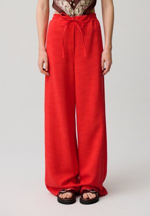 Mujer con pantalones sueltos rojos con cordón y sandalias negras, de pie contra un fondo liso gris claro.