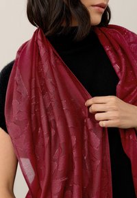 Translucent burgundy scarf with a floral pattern and shiny texture, draped over a black top, held by a hand at one edge.