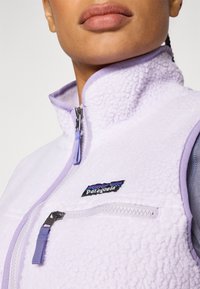 Gaiši purpursarkans flīsa veste ar augstu apkakli, ar rāvējslēdzēju priekšā un rāvējslēdzēju krūšu kabatu. Iespējas ietver zīmola etiķeti ar daudzkrāsainu kalnu dizainu.