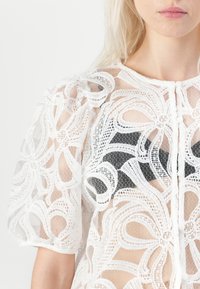 Haut en dentelle blanche semi-transparente avec un motif floral, des manches bouffantes, un col rond et une fermeture à boutons à l'avant, superposé sur une base sombre.