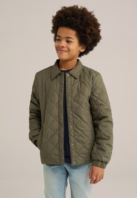 Quilted olive-green jacket featuring a button-up front, a collar, and elastic cuffs. Worn over a navy t-shirt and light blue jeans.