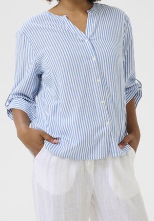 Person wearing a blue and white vertical striped button-up shirt with rolled sleeves and loose white linen pants, hands in pockets.