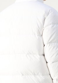Vit puffjacka med quiltat mönster, med hög krage och mjuk, slät textur. Synlig sömnad och rundade axelsömmar.