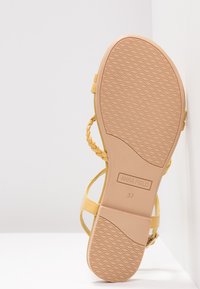 Sandale jaune avec des brides tressées, semelle plate et semelle extérieure en caoutchouc texturé. Présente le logo sur la semelle intérieure. Taille 37 indiquée au fond.