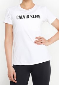 Mujer vistiendo una camiseta ajustada blanca de Calvin Klein y leggings negros, de pie con una mano en la cadera frente a un fondo blanco.