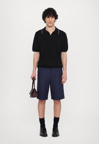 Polo shirt noir à côtes avec un col beige contrastant, associé à un short bleu marine. Le modèle tient un petit sac marron. Des chaussures et des chaussettes noires complètent le look.