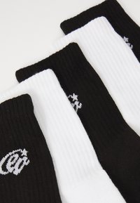 Drei Paare gerippte Socken: zwei schwarze und eine weiße, mit kontrastierenden farbigen Bündchen und kleinen, in Schwarz und Weiß gestickten Logos.