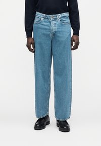 Hele sinised, laia säärega teksapüksid keskmise tõusuga vöökohaga, esitaskud ja julged õmblused. Sobitatud mustade kingadega.