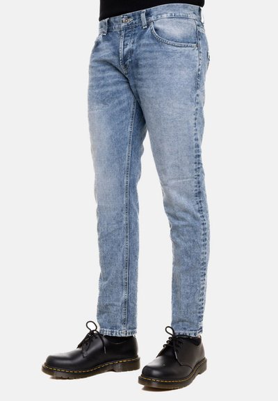 Jeans slim fit blu chiaro con un design sbiadito, quattro tasche e una chiusura con zip standard, abbinati a scarpe nere con lacci.