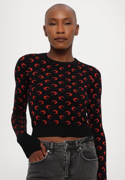 Černý zkrácený svetr s červeným vzorem půlměsíce, žebrovanými manžetami a kulatým výstřihem, doplněný šedými džíny. Hladká pletená textura.