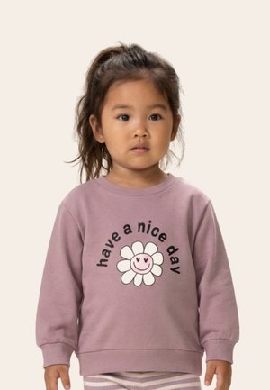 Jauna meitene ar tumšiem matiem astē, tērpusies mauves krāsas džemperī ar smaidīgu ziedu un uzrakstu "have a nice day".