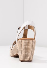 Białe sandały na koturnie z plecionym, teksturowanym obcasem, regulowanym paskiem i brązową wkładką. Projekt obejmuje otwarte boki i zamknięty tył.