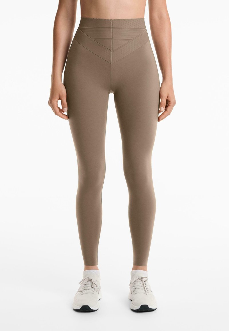 Hoge taille, naadloze leggings in een lichtbruine kleur met een gladde textuur en geometrische naaddetails. Gecombineerd met witte sportschoenen.