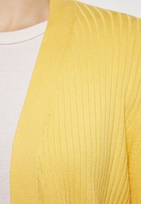 Cardigan galben cu dungi, având un material texturat și un design cu înaintare deschisă, purtat peste un top ajustat roz deschis.
