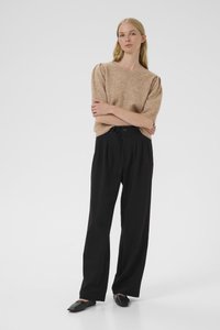 Pullover beige a maglia con maniche corte, abbinato a pantaloni neri sartoriali a vita alta. Il modello è in piedi con le braccia incrociate, indossando ballerine nere.