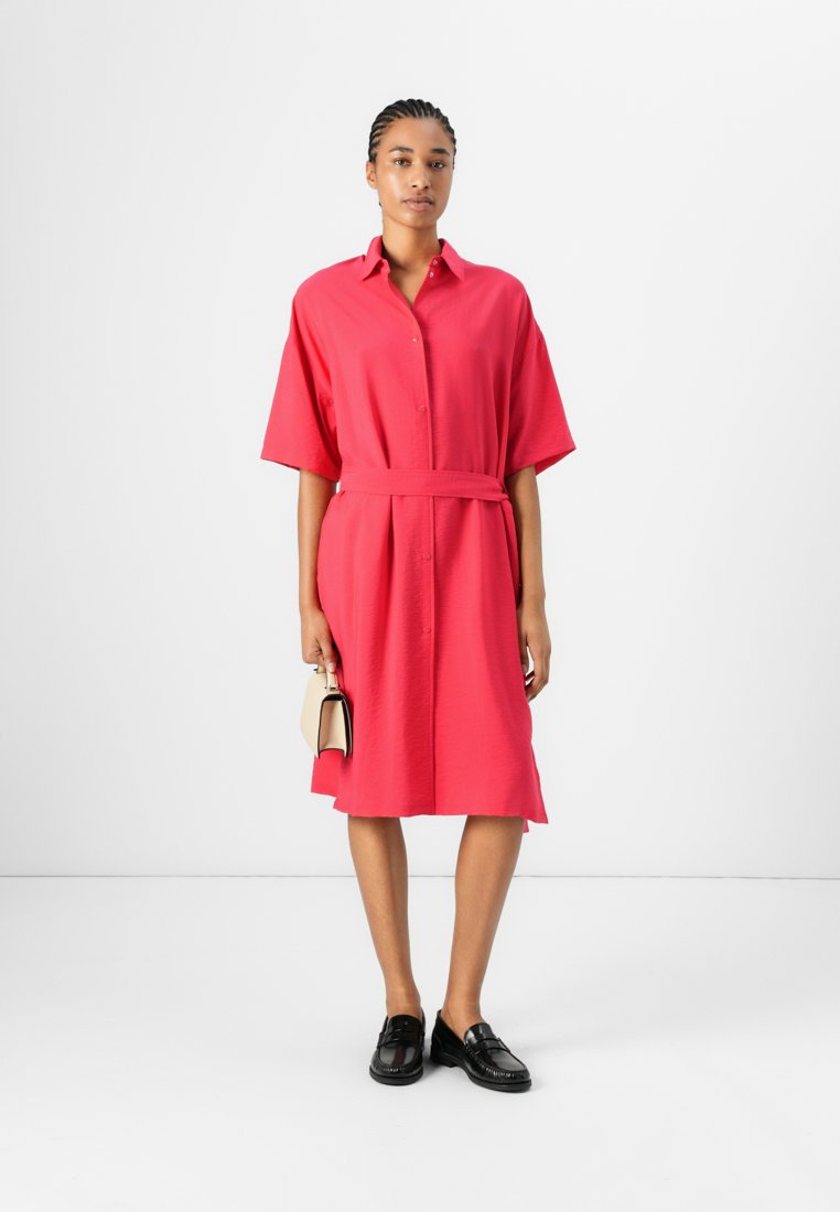 Robe chemise rose à manches courtes avec taille ceinturée, col et devant boutonné, associée à des mocassins noirs et un petit sac à main beige.