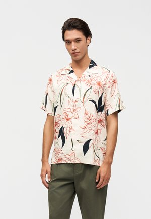 Mężczyzna w kremowej krótkiej koszuli na guziki z dużym czerwono-czarnym kwiatowym wzorem i oliwkowozielonych spodniach, stojący na białym tle.