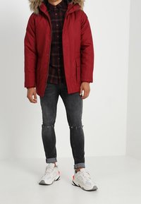 Abrigo de tela roja con capucha forrada de piel, combinado con una camisa a cuadros y jeans grises rasgados. Calzado con zapatillas deportivas blancas y grises.