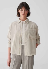 Beige corduroy shirt with vertical stripes, button closure, collar, and single chest pocket. Long sleeves with rolled cuffs.