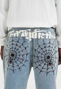 Lichtblauwe denim jeans met zwarte spinnenwebborduursels op de achterzakken, met een groot wit tekstontwerp over de tailleband.