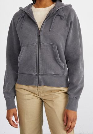 Personne portant un sweat à capuche gris zippé avec poches avant et un pantalon beige, debout devant un fond clair uni.