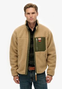 Kurtka z polaru w kolorze jasnobrązowym z zieloną kieszeni�ą na piersi, zamkiem błyskawicznym i naszywką marki outdoorowej. Tkanina o wyraźnej fakturze i luźnym kroju.